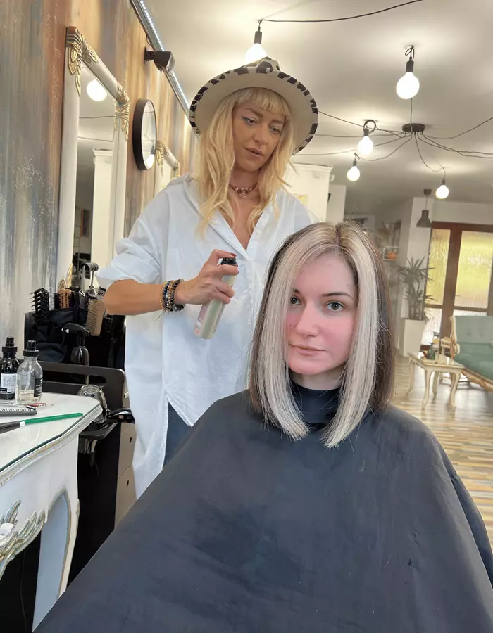 Stilistă lucrând în salon, cu grădina verde vizibilă prin ușile de sticlă din spate